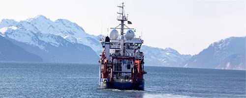 A research vessel is navigating through calm waters, with its vibrant red and blue hull contrasting against the serene backdrop of snow-capped mountains. The landscape showcases a clear sky with subtle tones of light, emphasizing the natural beauty of the environment.