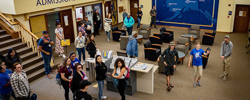 The scene captures a vibrant admission area at a university. A group of individuals is gathered around an information desk. In the background, a large blue wall mural outlines the campus locations in Alaska, stating, 'Welcome to Troth Yeddha' You belong here.'