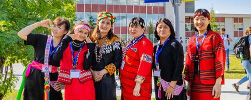 A group of women in traditional attire stands together outside at the University of Alaska Fairbanks. They are posing playfully, showcasing their colorful clothing, which includes intricate patterns and vibrant colors.