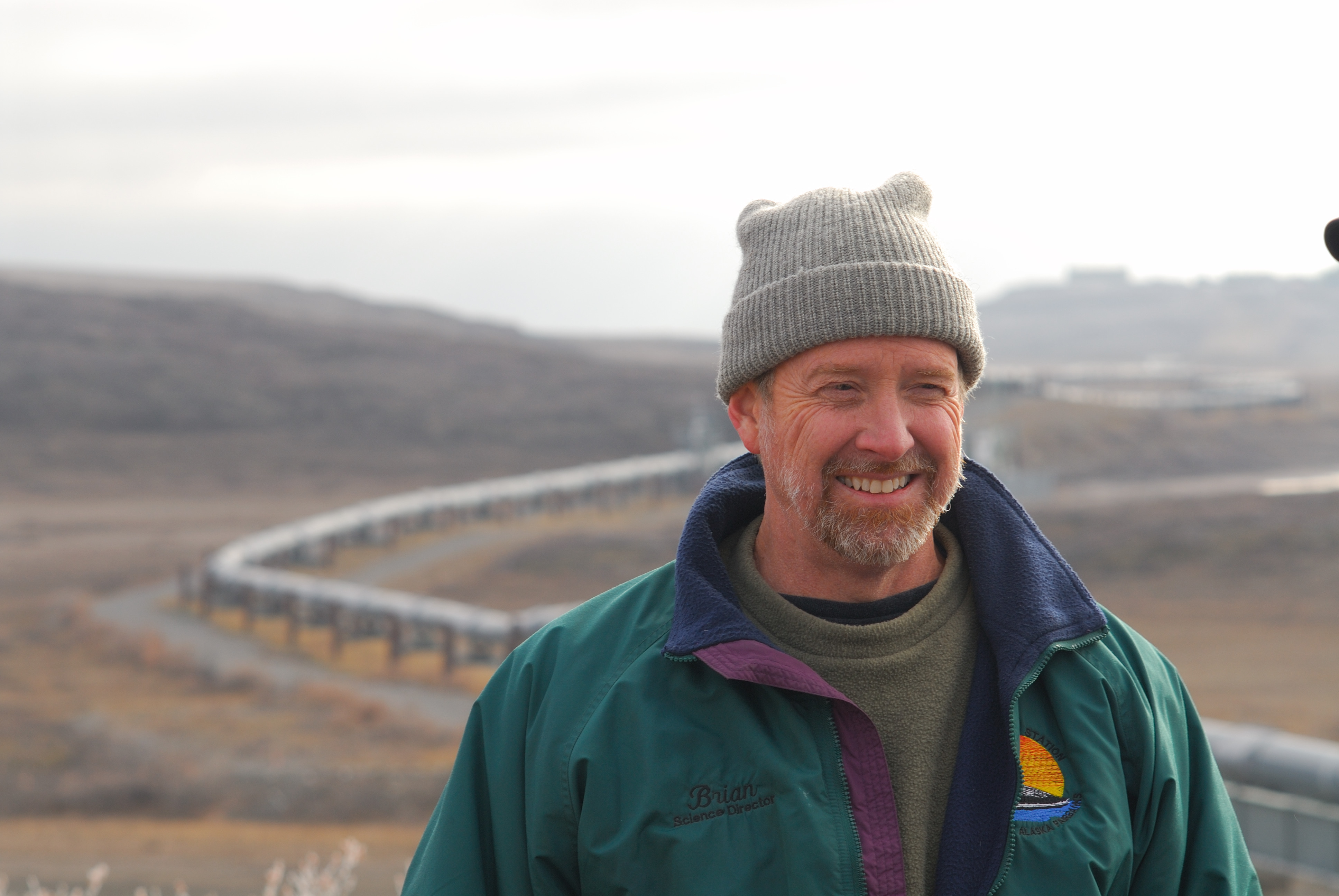 Head and shoulders photo of a man in a knit cap and light jacket in a fall landscape with the Alaska oil pipeline in the background.