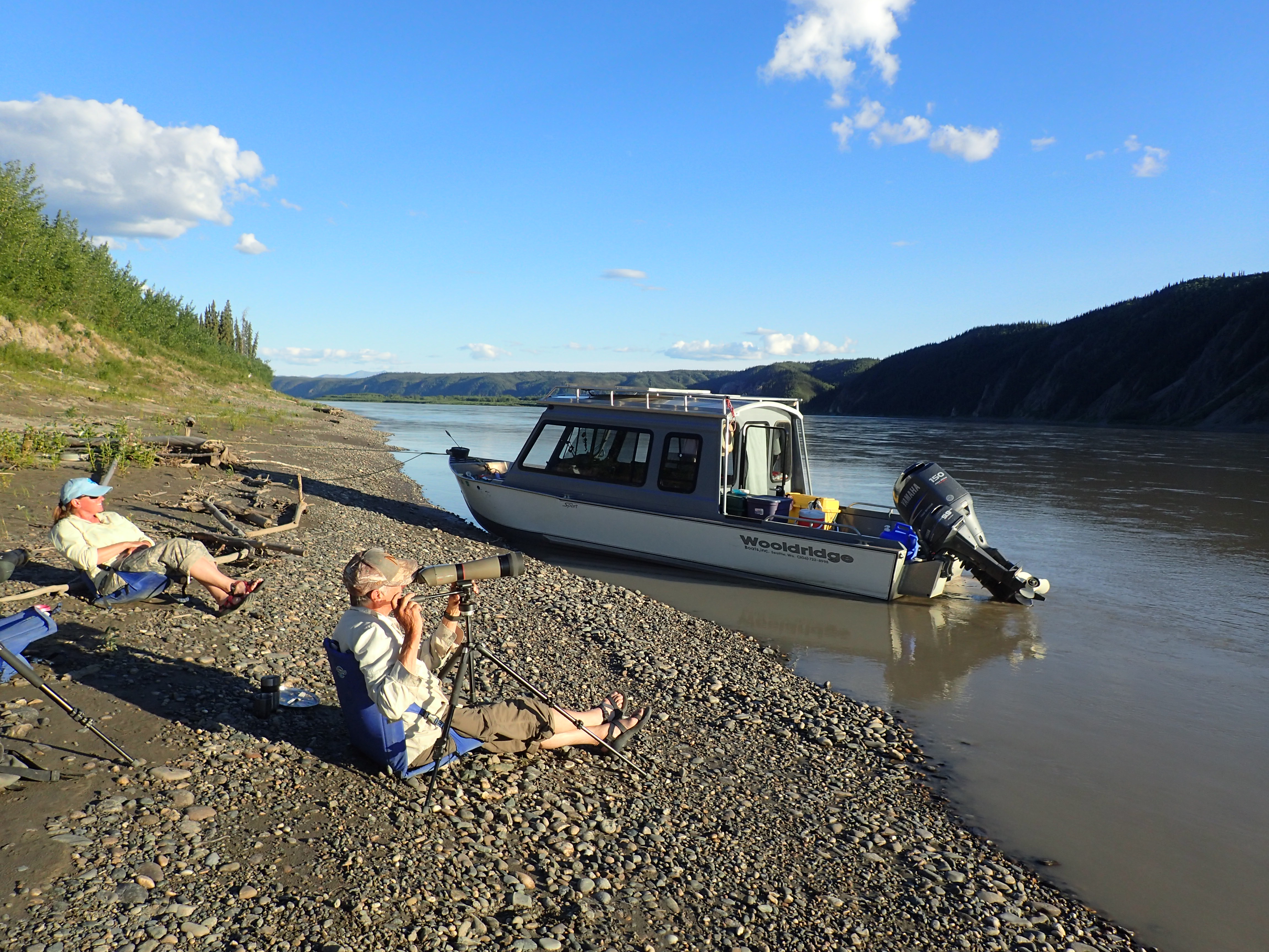 Two people sit on the pebbled riverbank with a small boat and outboard motor anchored nearby. One person looks through a long spotting scope on a tripod.