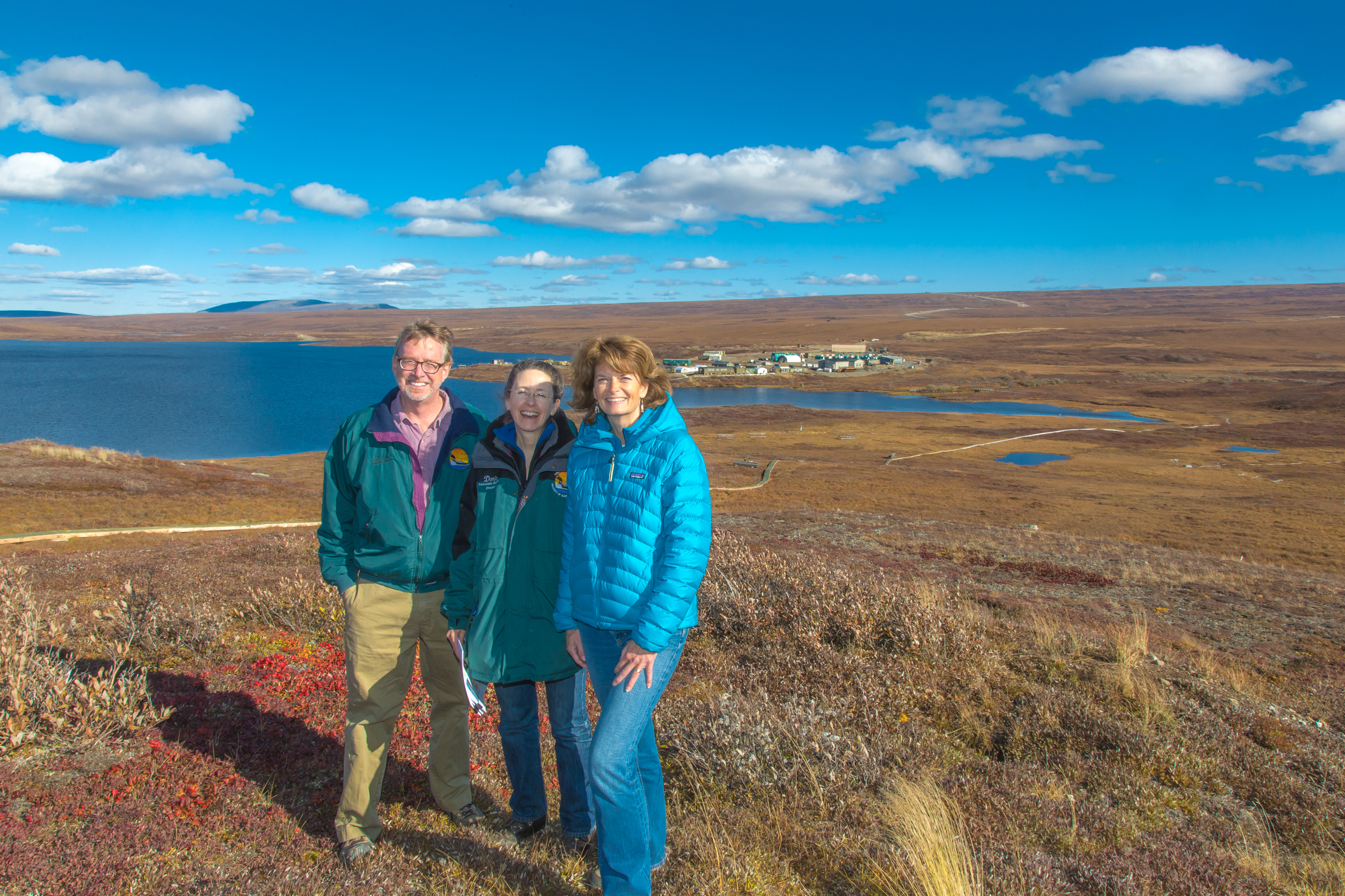 Brian Barnes, Donie Brett-Harte and Senator Lisa Murkowski standing in an autumn tundra landscape with Toolik Field Station in the distance.