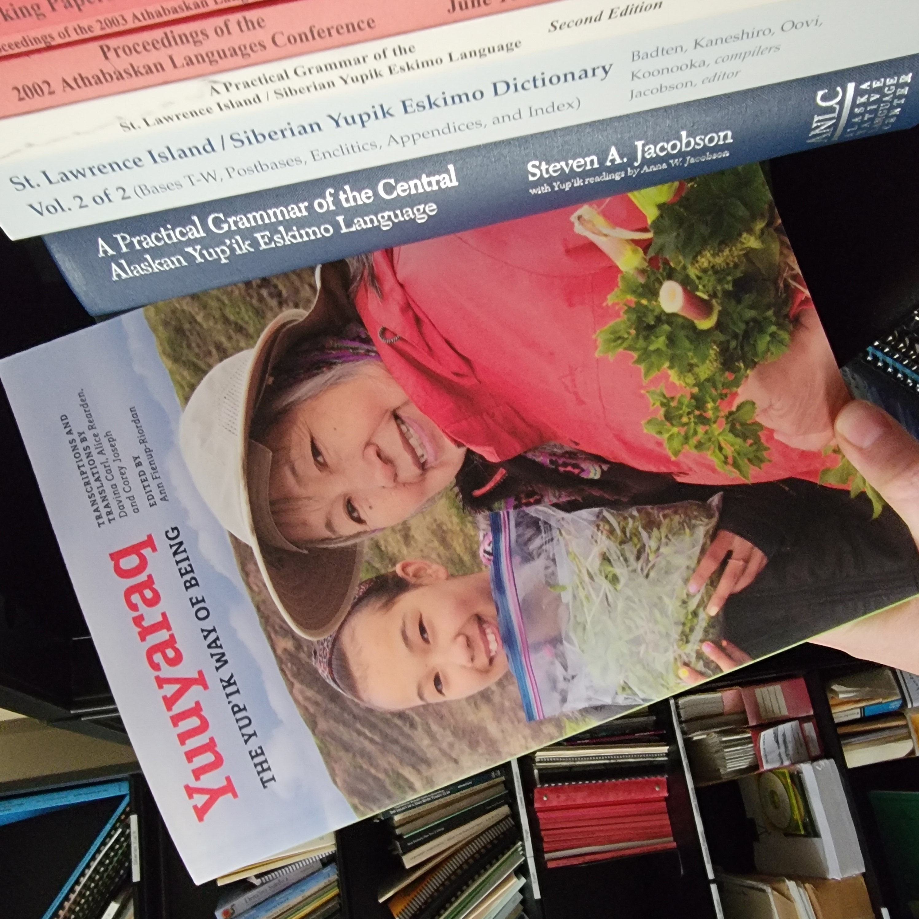 A person off-camera pulls a book with the title "Yuuyaraq, The Yup'ik Way of Being" from a bookshelf in the Alaska Native Language Center bookstore. The book cover features two smiling Alaska Native women and the greens they have foraged in a wild landscape.