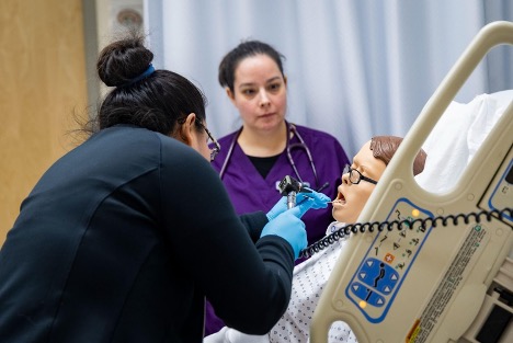 Two nursing students examine a mannequin patient lying in a hospital bed.