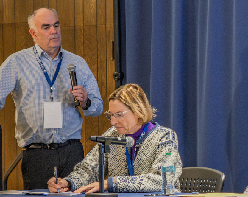 A man with a microphone stands to the left of a woman seated at a table taking notes with a pen and paper.