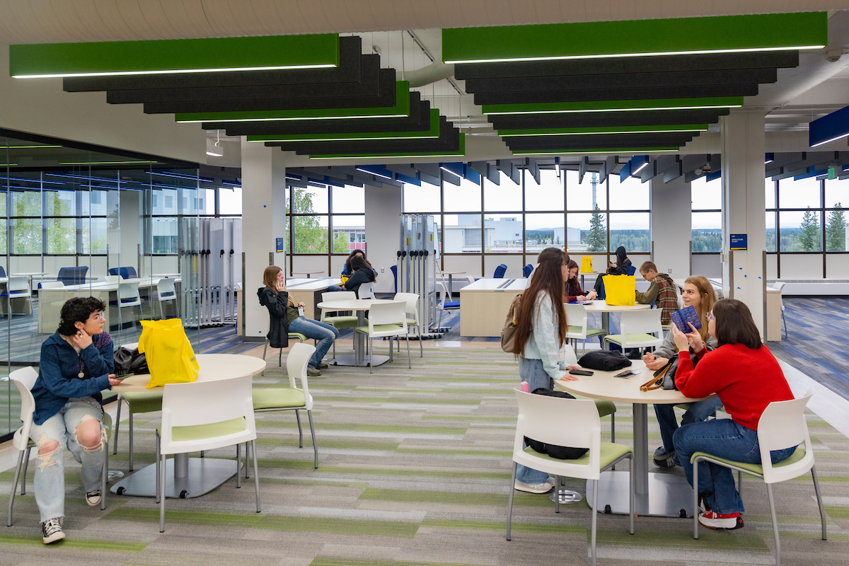 Groups of college students sit and interact at round tables in a large, airy, brightly lit space.