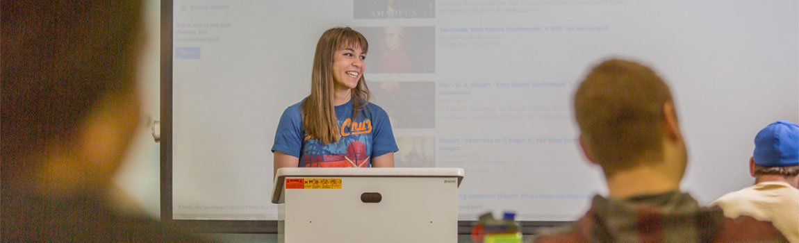A smiling student stands at a podium as with a projected presentation behind her. A group of students in the foreground look on.