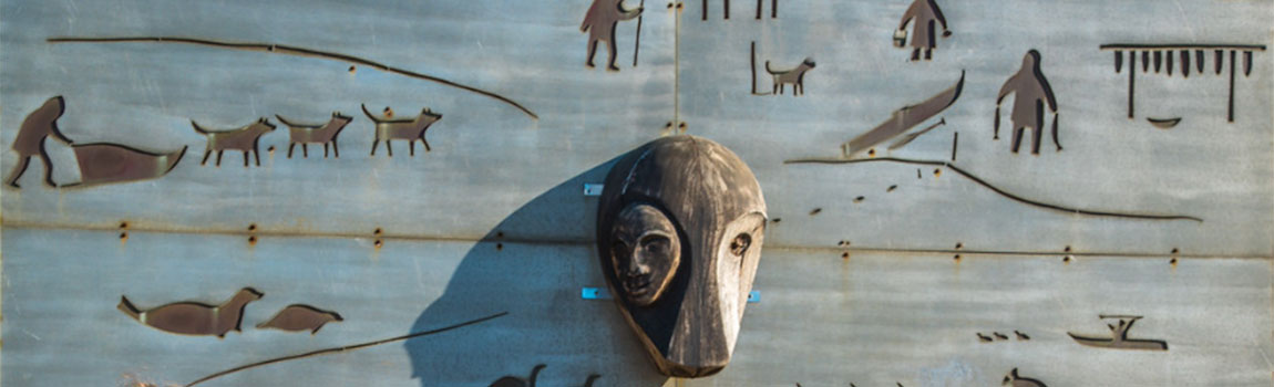 A sculpted mural featuring Alaska Native artwork silhouette depictions of dog sledding, seals, kayaking, drying salmon and a mask