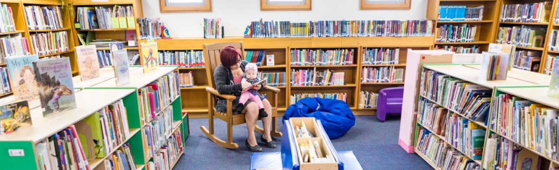 A woman sits in a rocking chair in a library reading to an infant sitting on her lap.
