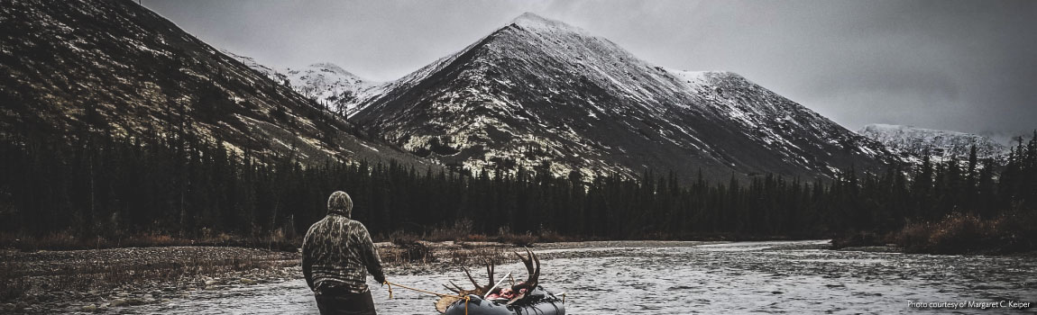 Photo of person pulling a raft with moose antlers down a river with mountains in the background.