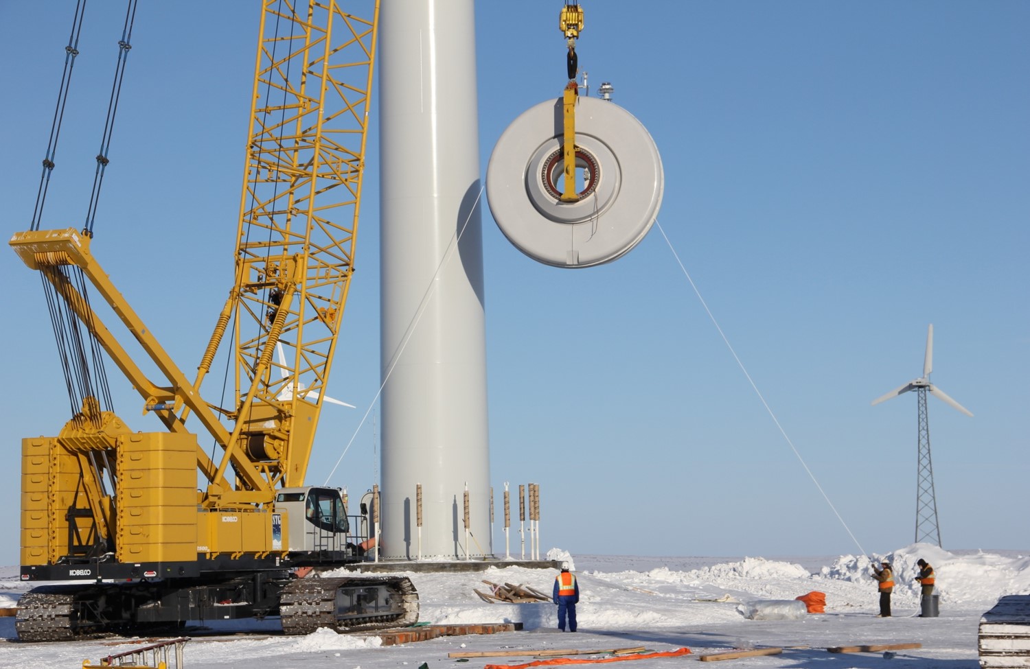 Large orange crane lifts round wind turbine part during winter installation. Small turbine visible in background.