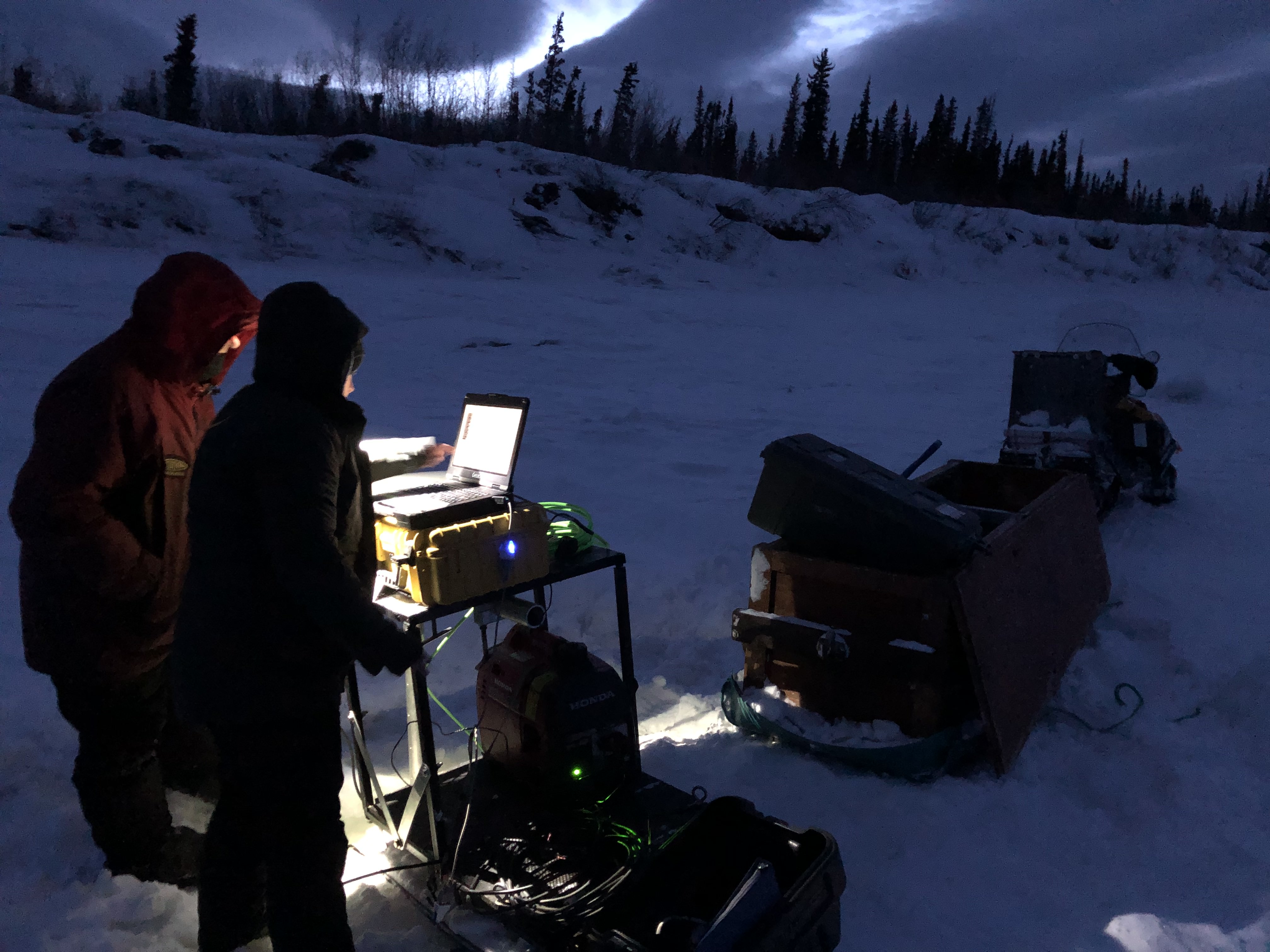 Two researchers stand in front of equipment in a dark winter scene illuminated by headlamps