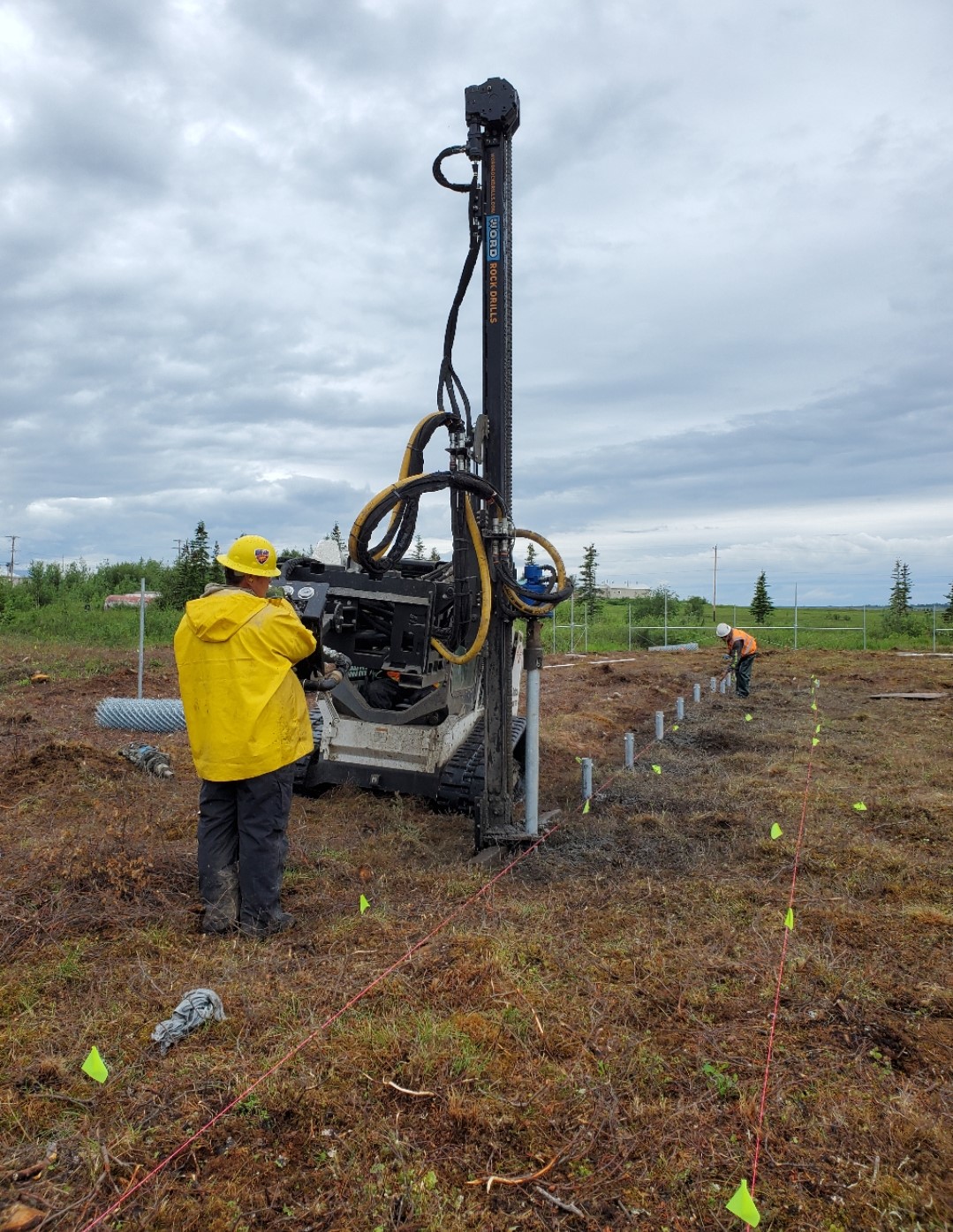 Man in yellow jacket stands on the tundra by a large drilling machine