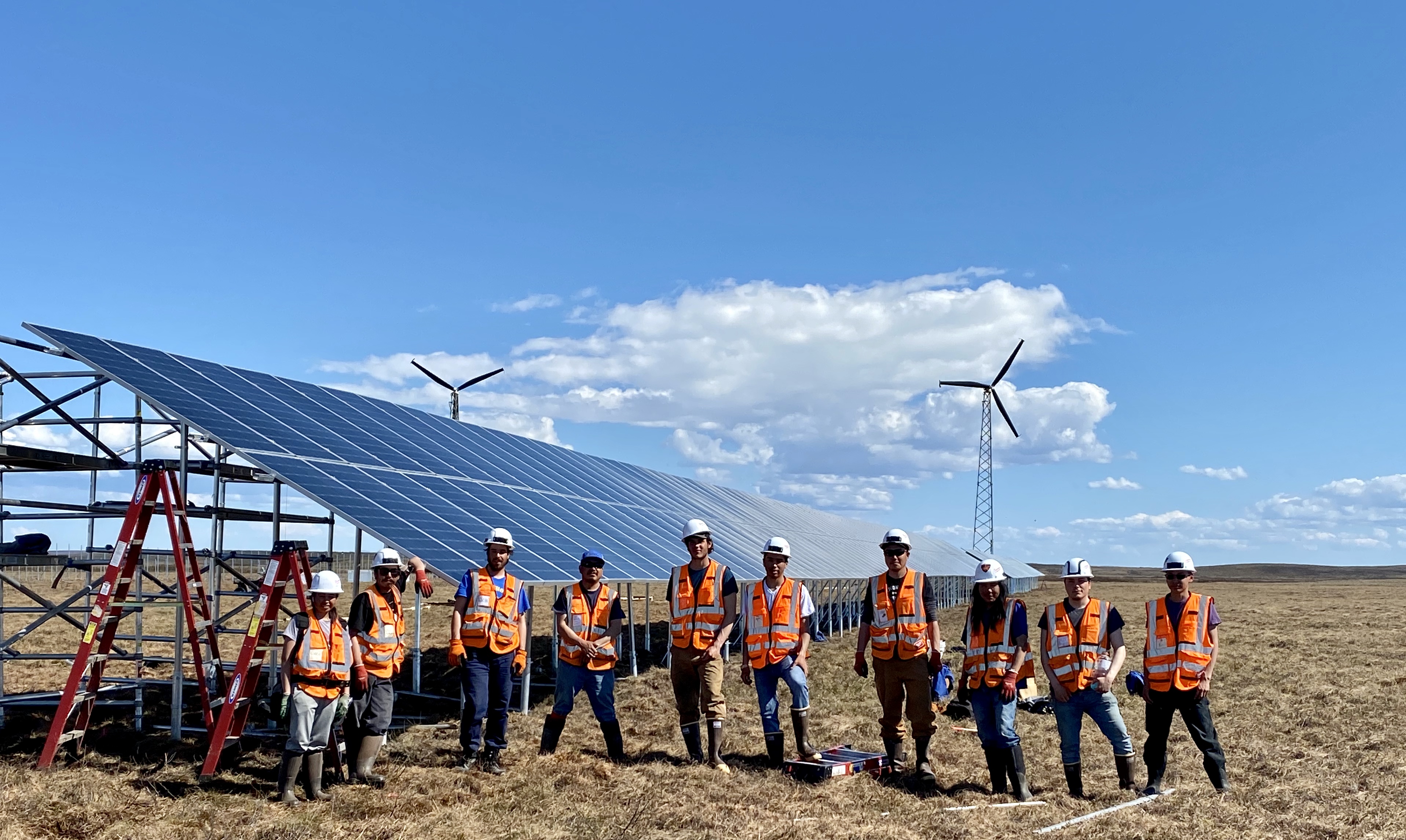 Photo of many people wearing vests and hard hats in front of solar panels on a sunny day. They look proud of their work.