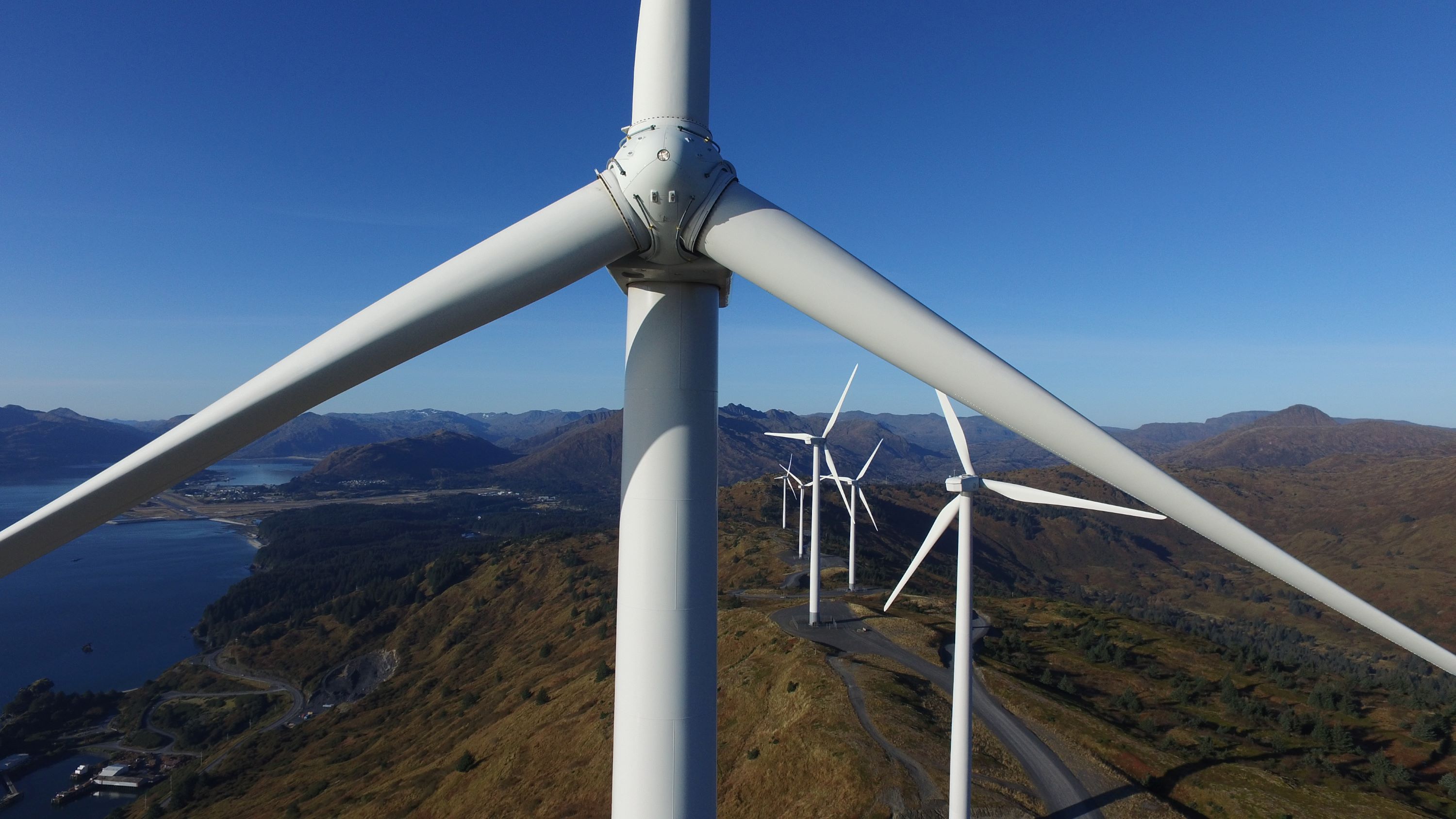 View of wind turbines on a clear summer day in a mountainous landscape. Water visible on left. 