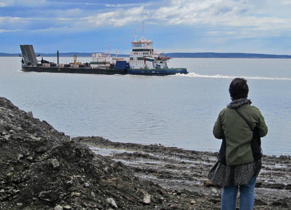 woman stands on a rocky beach and watches a large fuel barge pass by on the water.