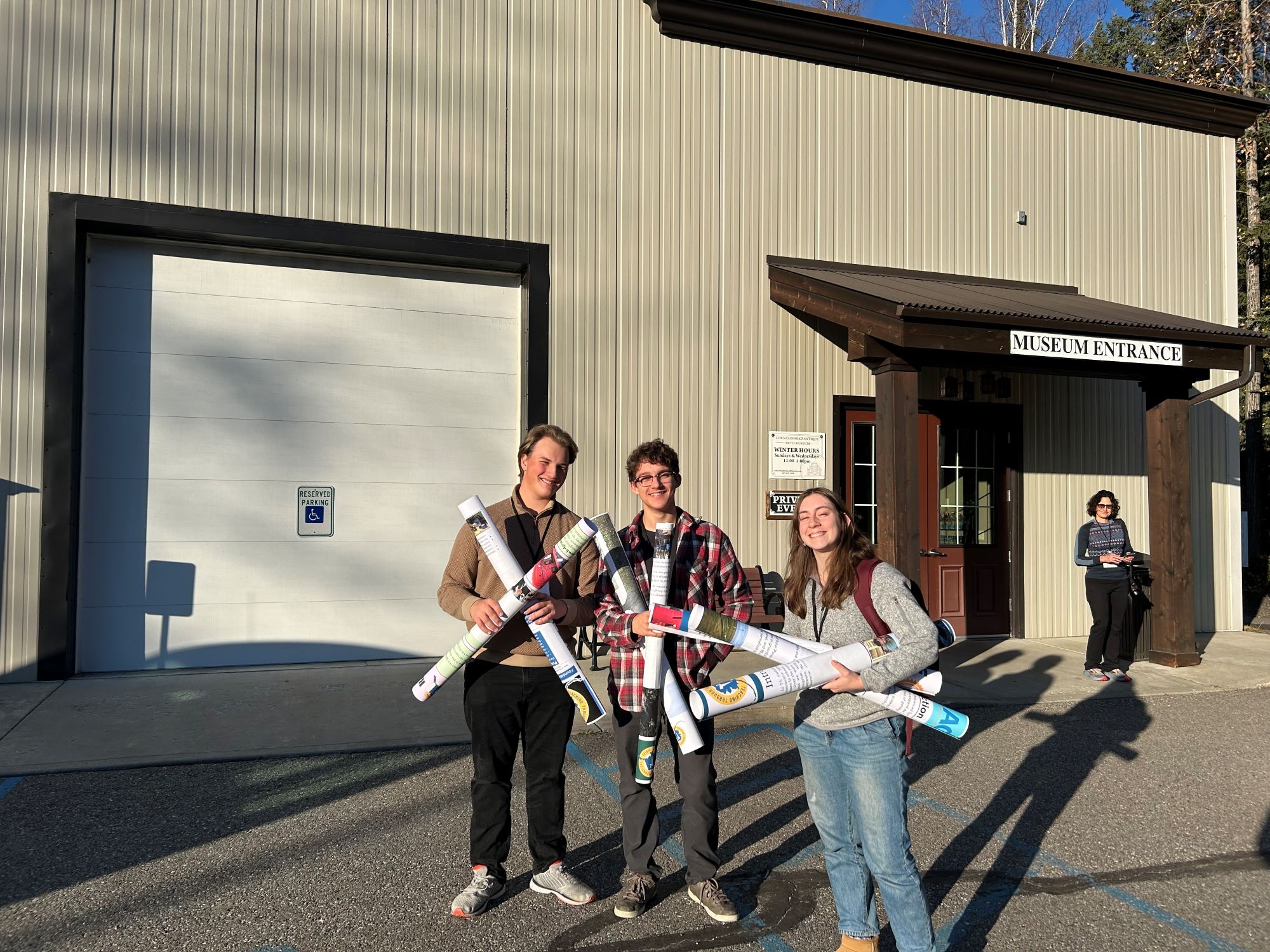 Students stand outside a museum entrance holding rolled up posters and smiling.