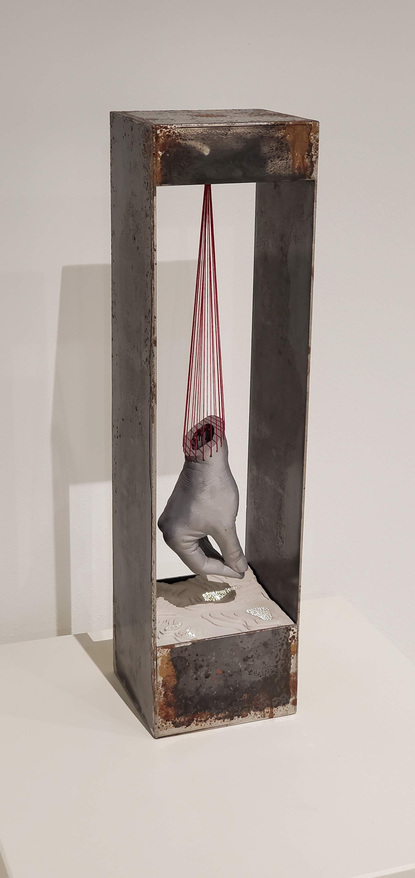 cast concrete hand hanging from red thread in an aluminum box with a topographic map at the base,  image courtesy of the artist