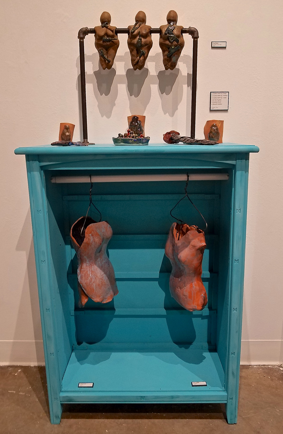 Blue dresser with an open front where we see two ceramic torsos suspended. Ceramic figures are on top. Image courtesy of Olena Ellis