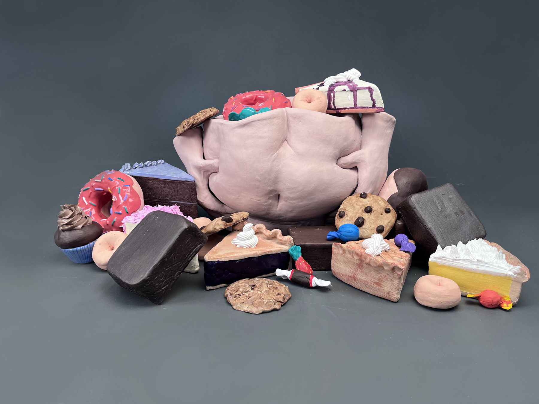A ceramic sculpture of hands grasping a belly surrounded by ceramic desserts, courtesy of the artist