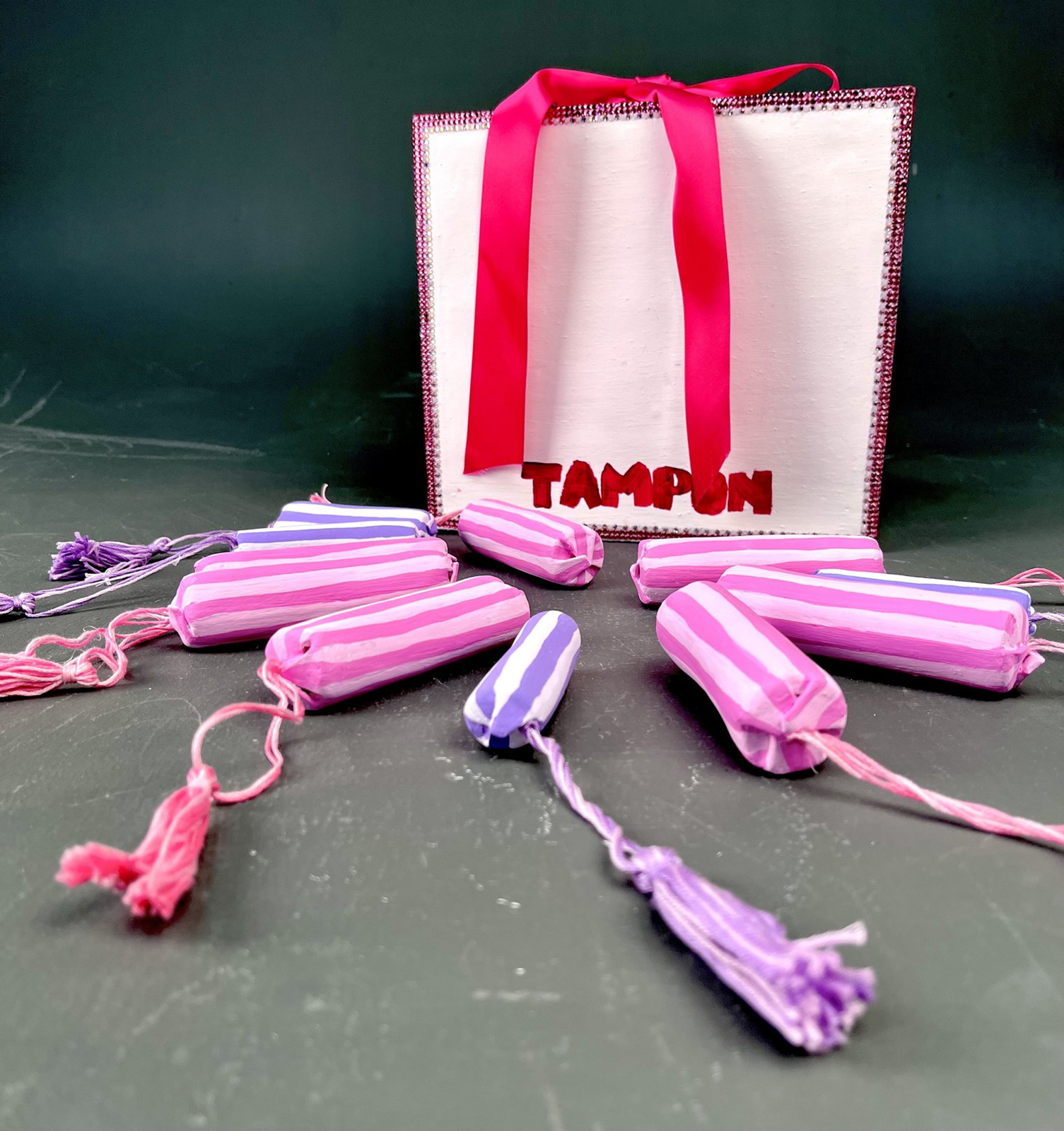A bag with the word "Tampax" and several ceramic and fabric tampons on the table in front of it, courtesy of the artist