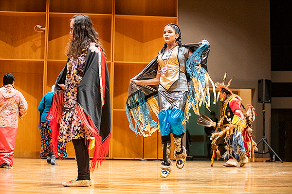A girl wearing a fringed cape and moccasins jumps while dancing on stage at the 2019 Festival of Native Arts. UAF photo by JR Ancheta.