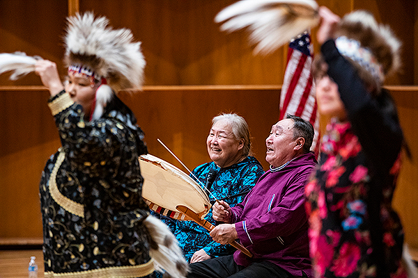 A man sitting next to a woman drums while two dancers perform at the 2019 Festival of Native Arts. UAF photo by JR Ancheta.