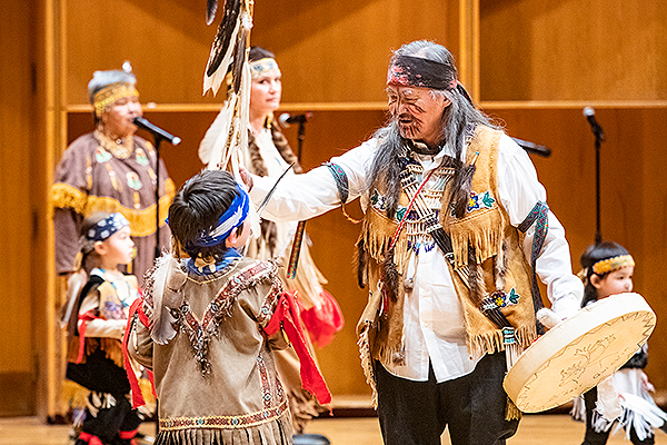 An older man in traditional dress and holding a drum and feather-covered spear interacts with a young boy on stage during the 2019 Festival of Native Arts. UAF photo by JR Ancheta.
