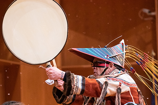 A man wearing a traditional painted and decorated wooden visor hat plays a drum during the 2023 Festival of Native Arts.