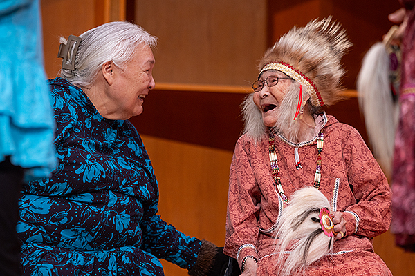 Two elderly women in traditional attire share a light-hearted moment during day two of the 2024 Festival of Native Arts. UAF photo by Eric Engman.