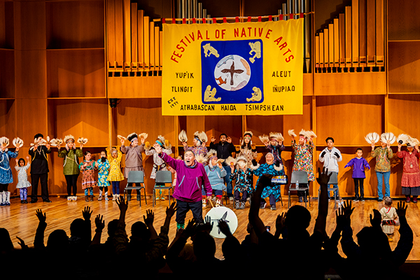 Performers of all ages take the stage on day three of the 2024 Festival of Native Arts. They engage the audience with raised hands, showcasing a rich display of cultural attire and traditional dance. UAF photo by Leif Van Cise.
