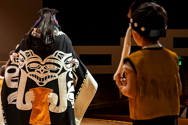 A person in traditional regalia, including a black and white cloak, performs on stage while a child watches during day three of the 2024 Festival of Native Arts. UAF photo by Leif Van Cise.