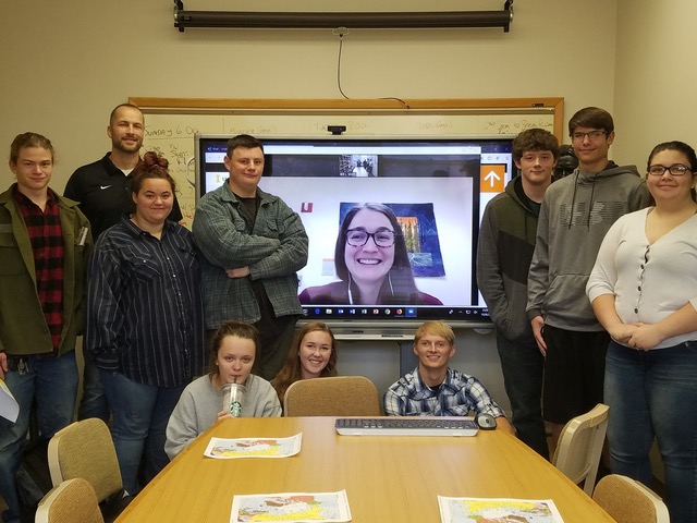 A group of people stand around a videoconferencing screen with a woman's face on it.