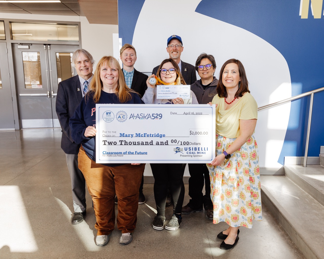 a teacher and student hold up checks posing with the UAF chancellor and four other staff and donors