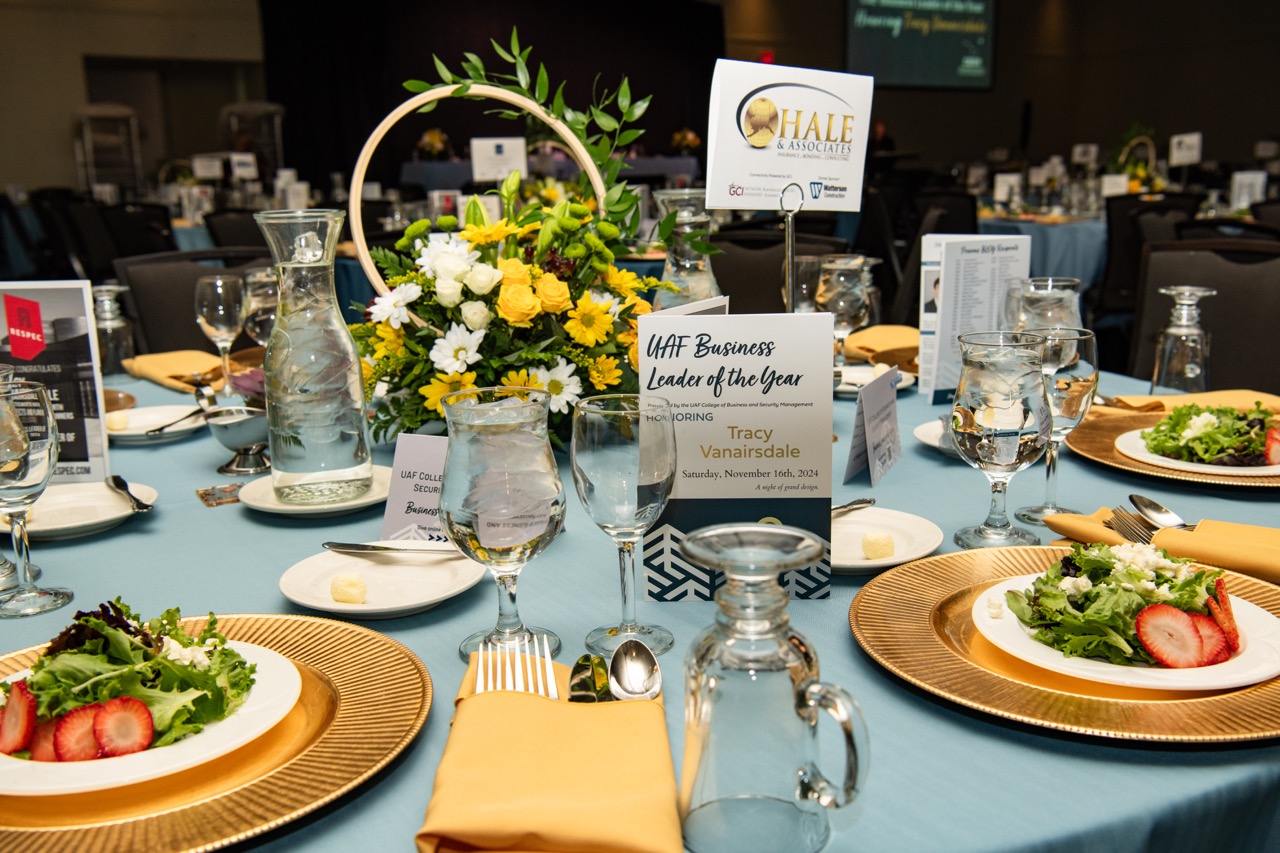 a table at an event set with a strawberry salad, silverware, glasses, and an event program reading "UAF Business Leader of the Year, November 16, 2024" and a sign for Hale & Associates