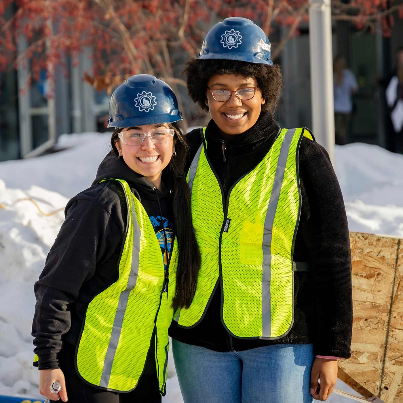 Two female students wearing reflective vests and hard hats are looking at the camera and smiling.