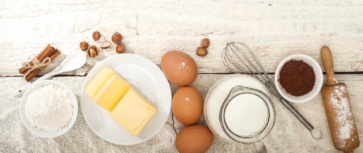 A wooden table displaying various baking ingredients, including flour, sugar, eggs, and butter