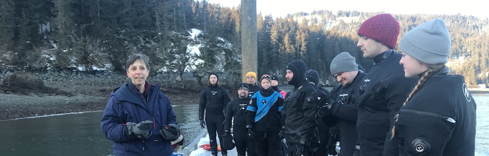 Photo of diving instructor Brenda Konar and her students, standing on the dock of the Kasitsna Bay Lab location, with snowy hills in the background