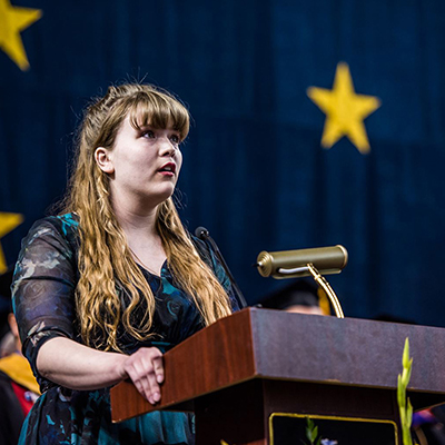 A student addresses an audience from a podium with the flag of Alaska hanging in the background