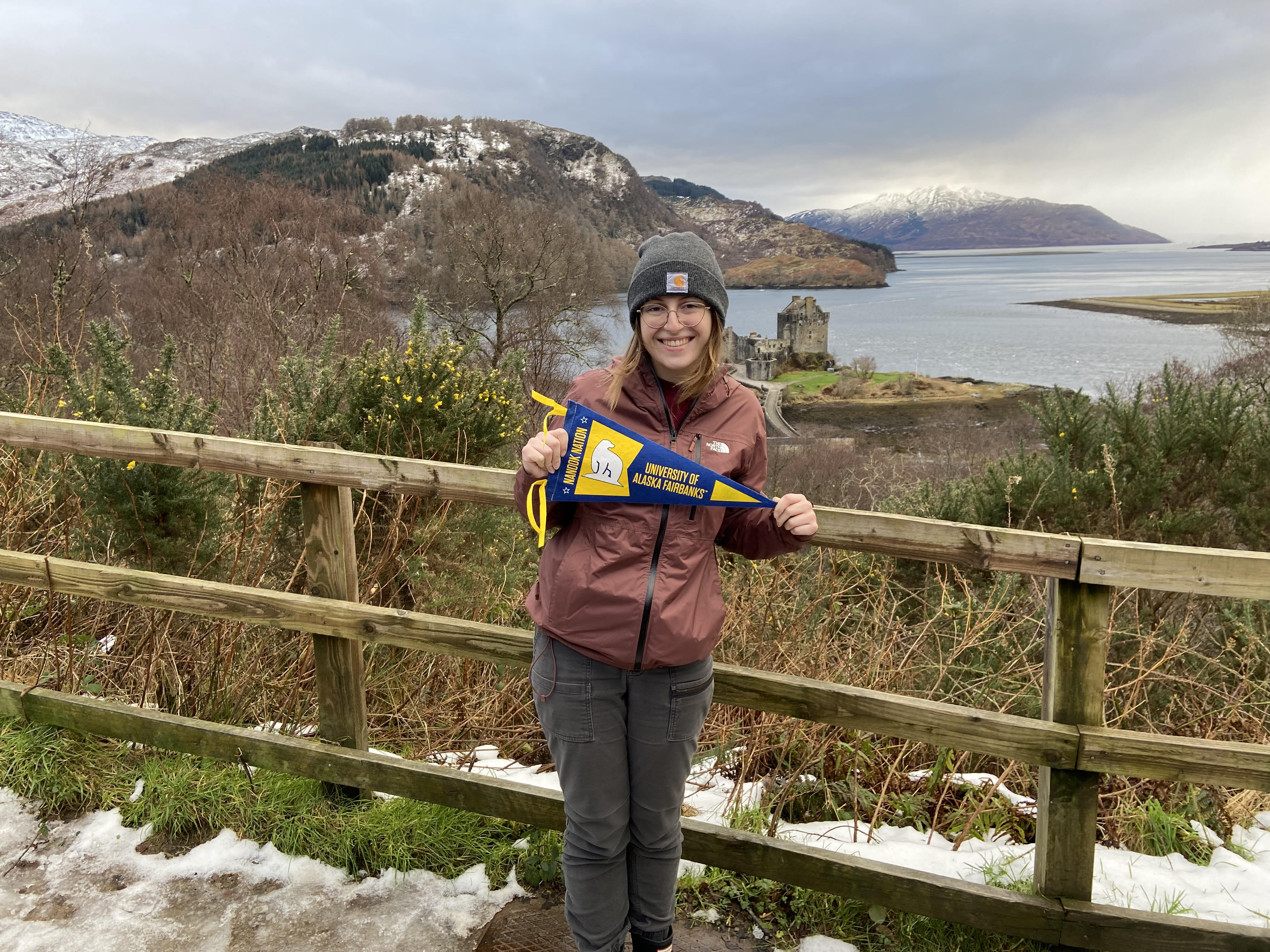 Study away student holding UAF flag, standing in front of a lake surrounded by mountains.