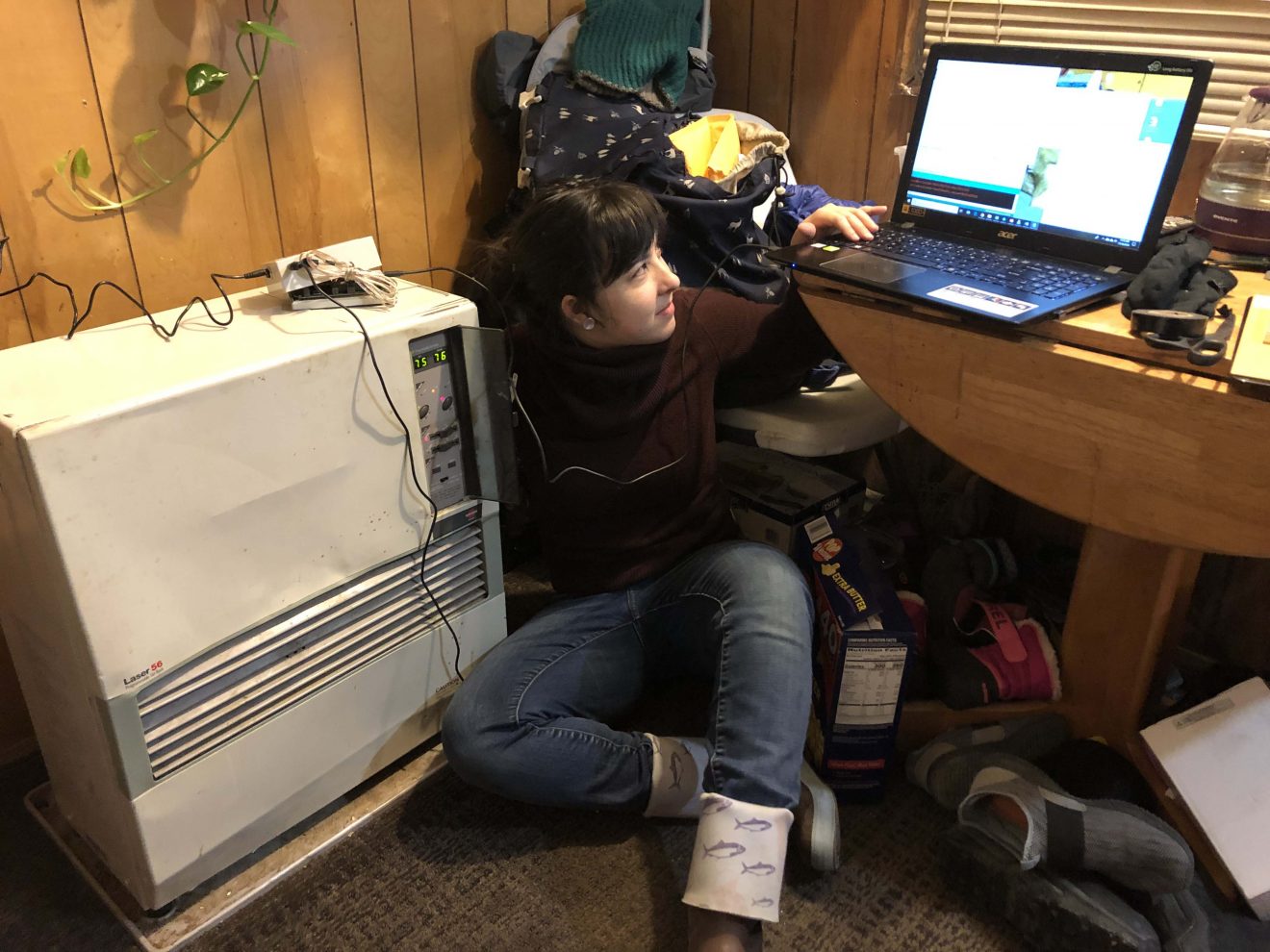 A woman sits on the floor between a heating unit and a desk. She is looking at a laptop on the desk.