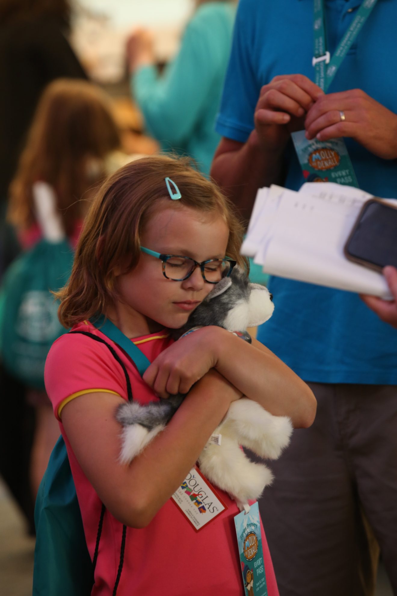 A young girl closes her eyes and hugs a stuffed dog. She stands near two adults and another child. Everyone is wearing Molly of Denali backpacks or lanyards.