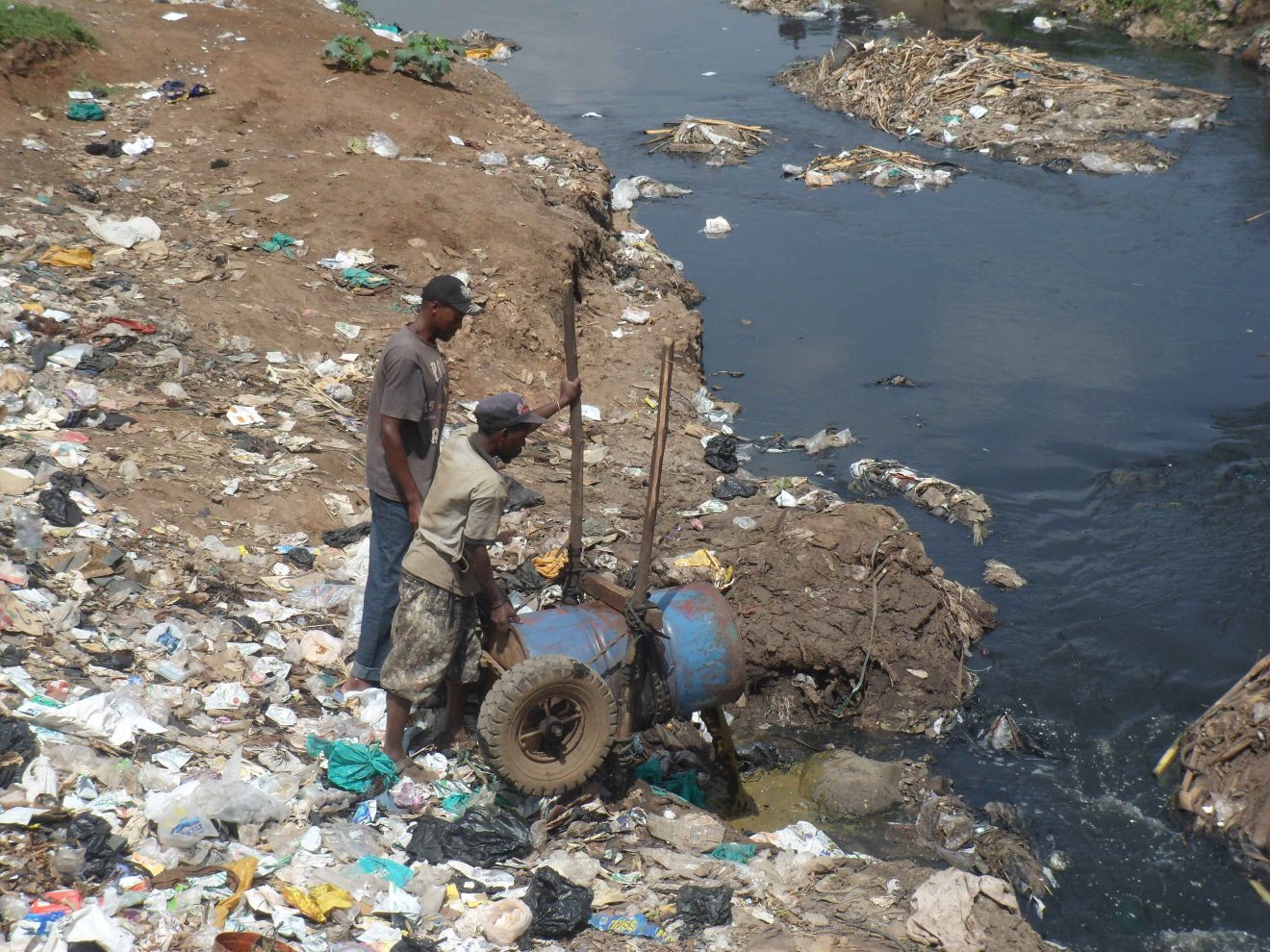 Two men pour a barrel of sludge into a trash-strewn waterway.