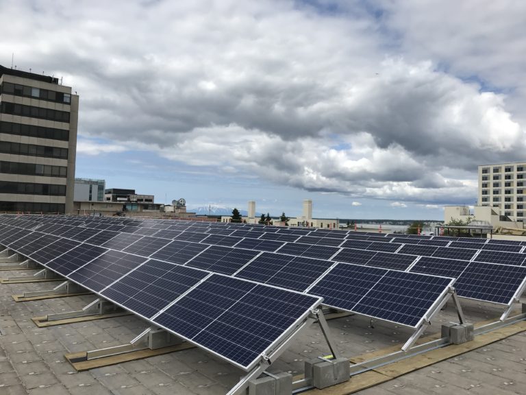 Six long rows of solar panels on a rooftop in Anchorage. Several other taller buildings surround.