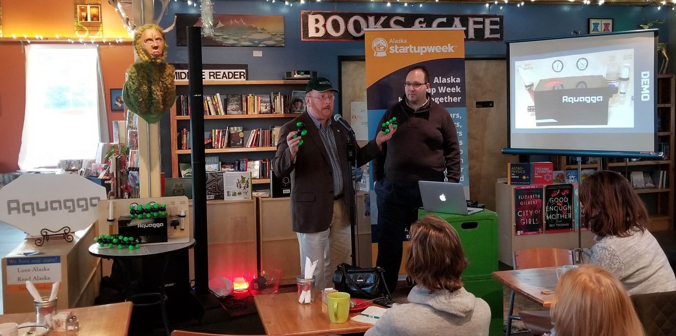 Two men stand in a bookstore. One man holds a multipronged green object in each hand. A screen is set up beside them. Three people sit at desks looking at them.