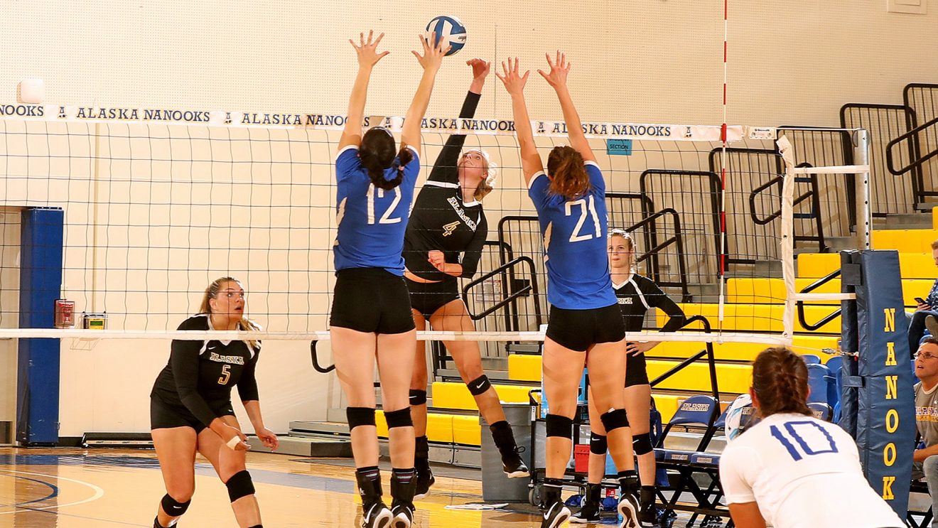 Three volleyball players go up to the net while three others look on.