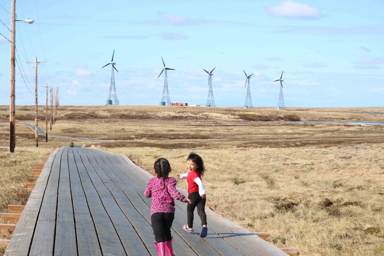 Two children run down a boardwalk laid across the tundra. Five wind turbines stand in the distance.