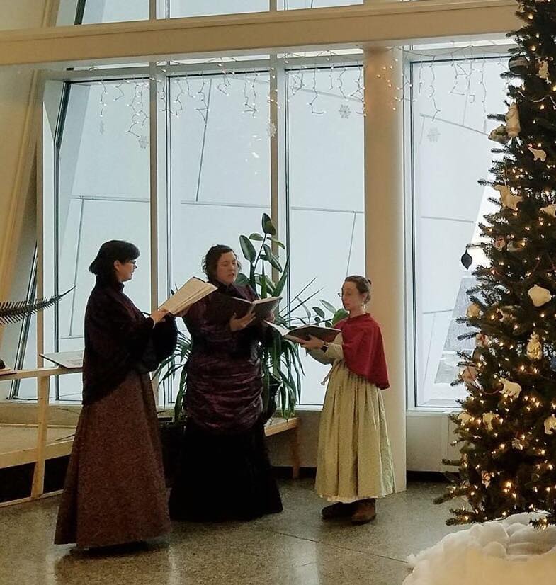 Three women in Victorian dress sing beside a Christmas tree.