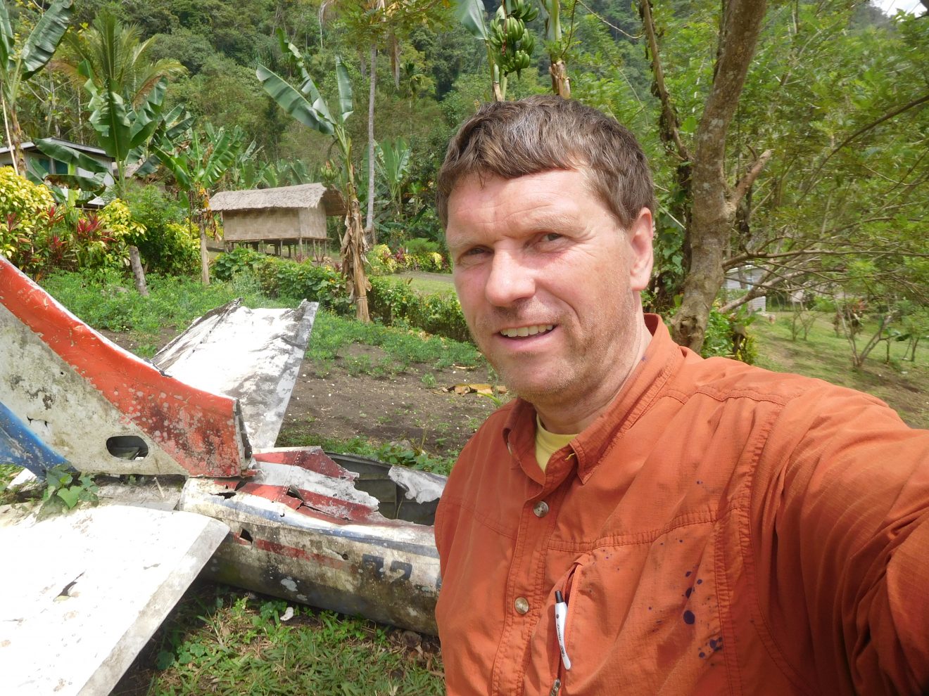 Selfie of man standing in green area (jungle?) with a crashed plane behind him.