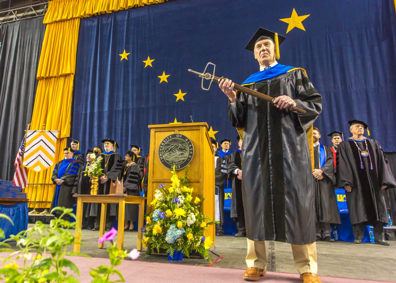 Stephen Jewett stands onstage at commencement holding the mace. He is dressed in cap and gown. The other members of the stage party, also in cap and gown, are behind him. A giant Alaska flag hangs in the background.